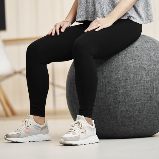 Frau sitzt auf grauem Design-Sitzball, Knie leicht unter Hüfthöhe, Füße flach am Boden