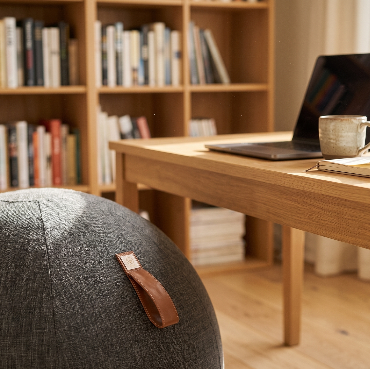 Treasy Sitzball in Anthrazit vor einem Holzschreibtisch mit Laptop, Kaffeetasse und Bücherregal im Hintergrund