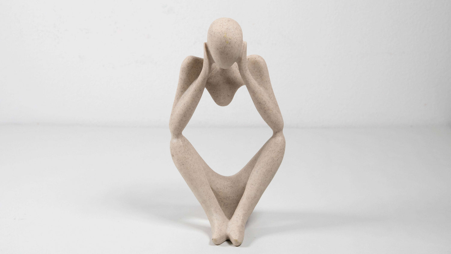 Eine minimalistische Figur aus Kunstharz sitzt mit angewinkelten Beinen und stützt den Kopf in die Hände. Die glatte, hellbeige Oberfläche und die abstrahierte Form verleihen ihr eine moderne, ruhige und nachdenkliche Ausstrahlung.