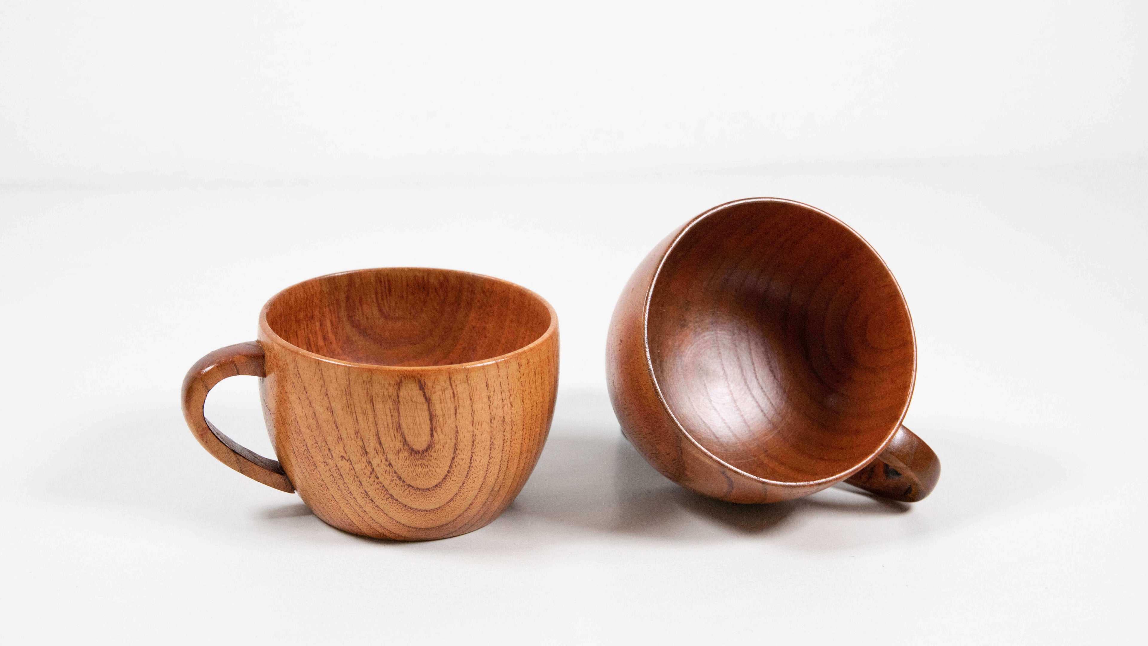 Zwei handgefertigte Teetassen aus Jujube-Holz auf weißem Hintergrund. Eine Tasse steht aufrecht, die andere liegt seitlich und zeigt ihre glatte, polierte Innenfläche mit feiner Holzmaserung.