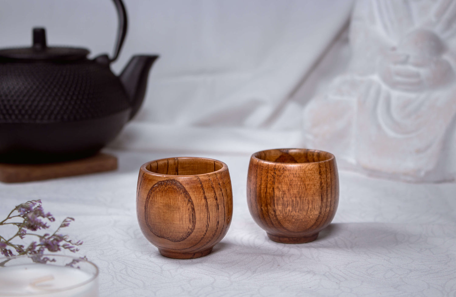 Das Foto zeigt zwei handgefertigte Teebecher aus Jujube-Holz, arrangiert in einem ruhigen, zen-inspirierten Setting. Im Hintergrund steht eine schwarze Gusseisen-Teekanne, flankiert von einer Buddha-Figur und dezenten Deko-Elementen. Die warme Holzstruktur der Becher hebt sich wunderschön vom weißen Untergrund ab und vermittelt Natürlichkeit, Achtsamkeit und japanische Teekultur – perfekt für eine Produktpräsentation mit Fokus auf Ruhe und Handwerk.