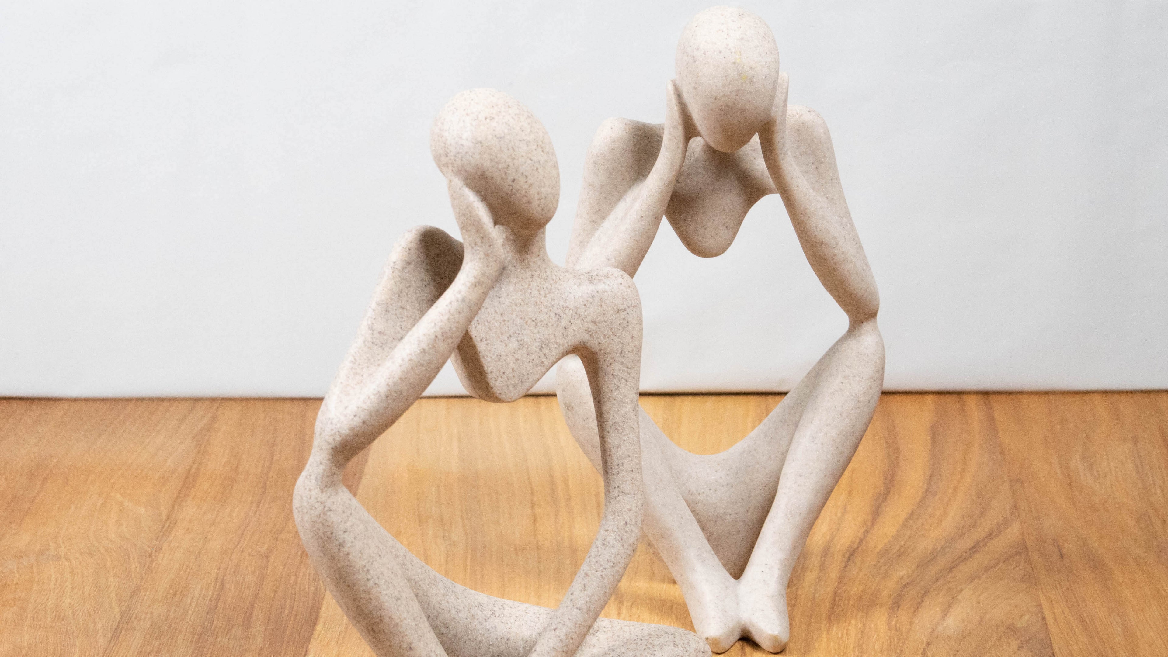 Zwei moderne Figuren aus Kunstharz sitzen auf einer Holzoberfläche in nachdenklicher Pose. Die abstrakten Formen mit glatter, steinähnlicher Oberfläche in hellem Beigeton verleihen der Szene eine ruhige, minimalistische Ästhetik.