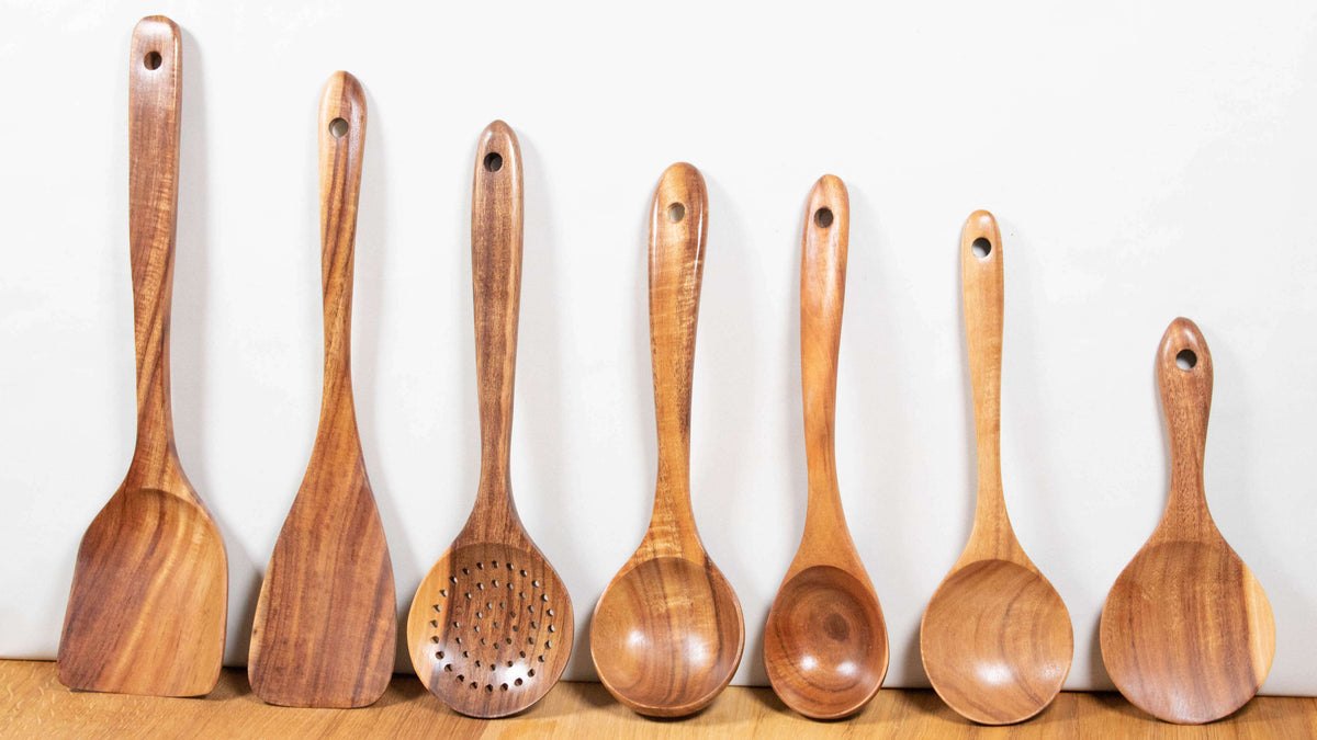 Sieben Kochutensilien aus Akazienholz nebeneinander an einer Wand: Wokspatel, Pfannenwender, Schaumkelle, Kellen und Reislöffel
